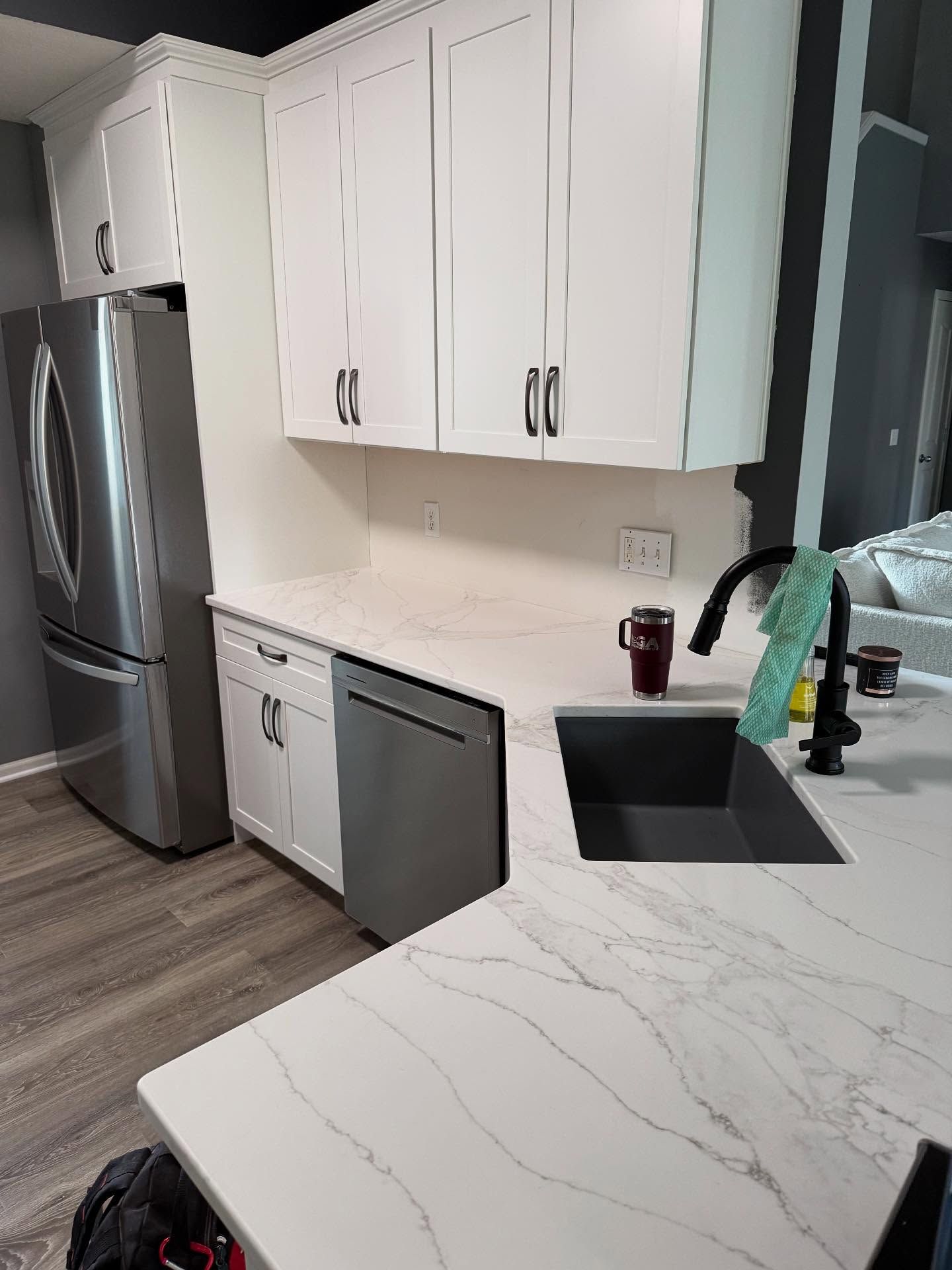 Modern white kitchen with stainless steel fridge, marble-look counters, black sink, and open dishwasher.