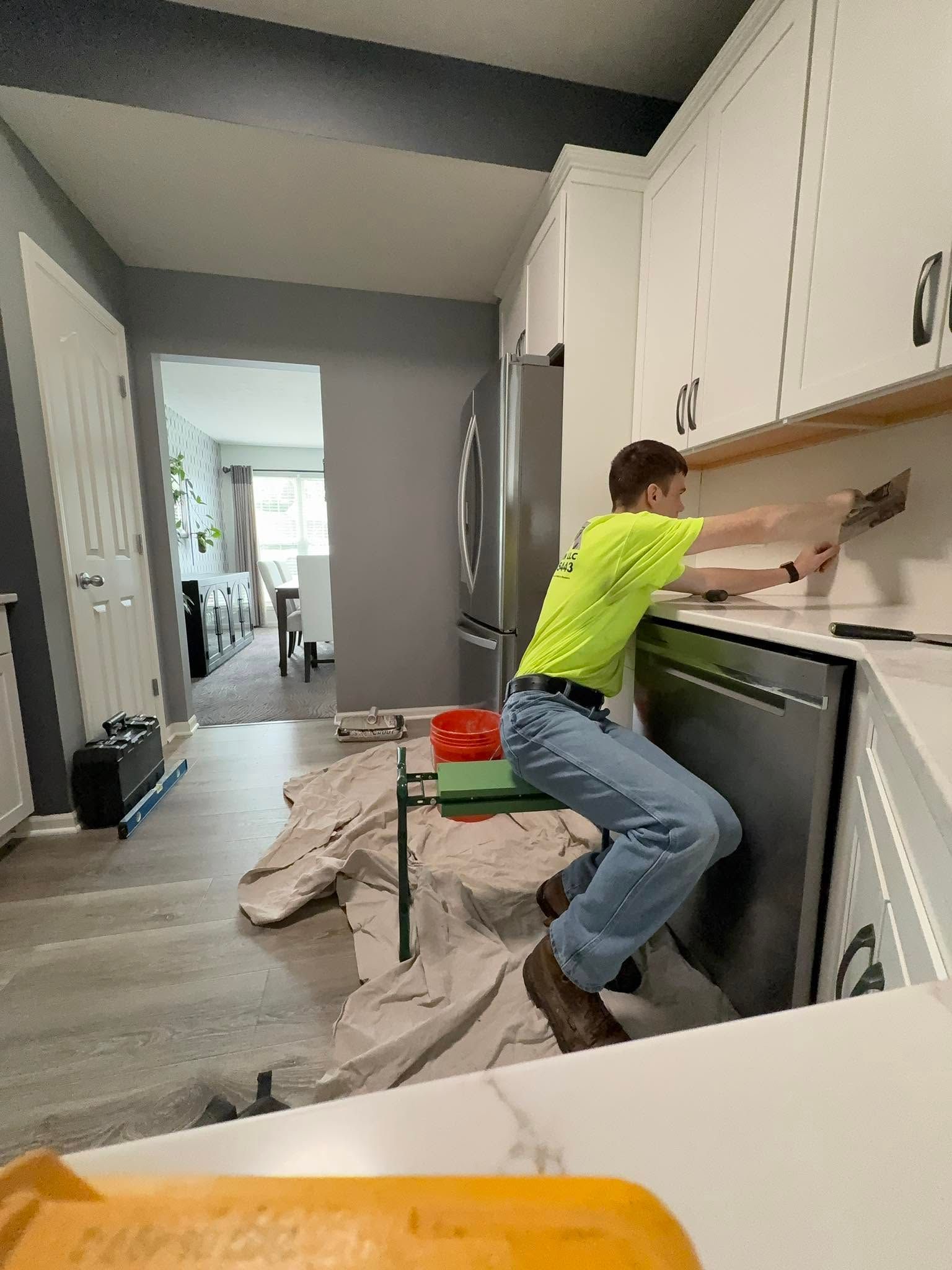 Person installing cabinetry in a white kitchen, kneeling on a drop cloth with tools and cabinets nearby.