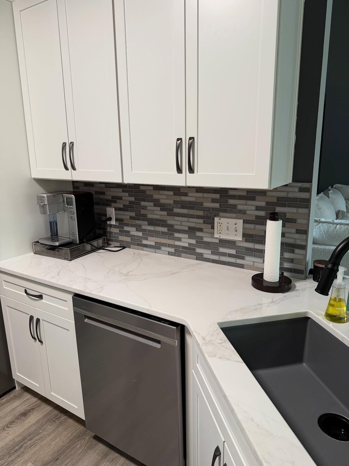 Modern white kitchen with gray backsplash, stainless dishwasher, sink, coffee maker, and soap on countertop