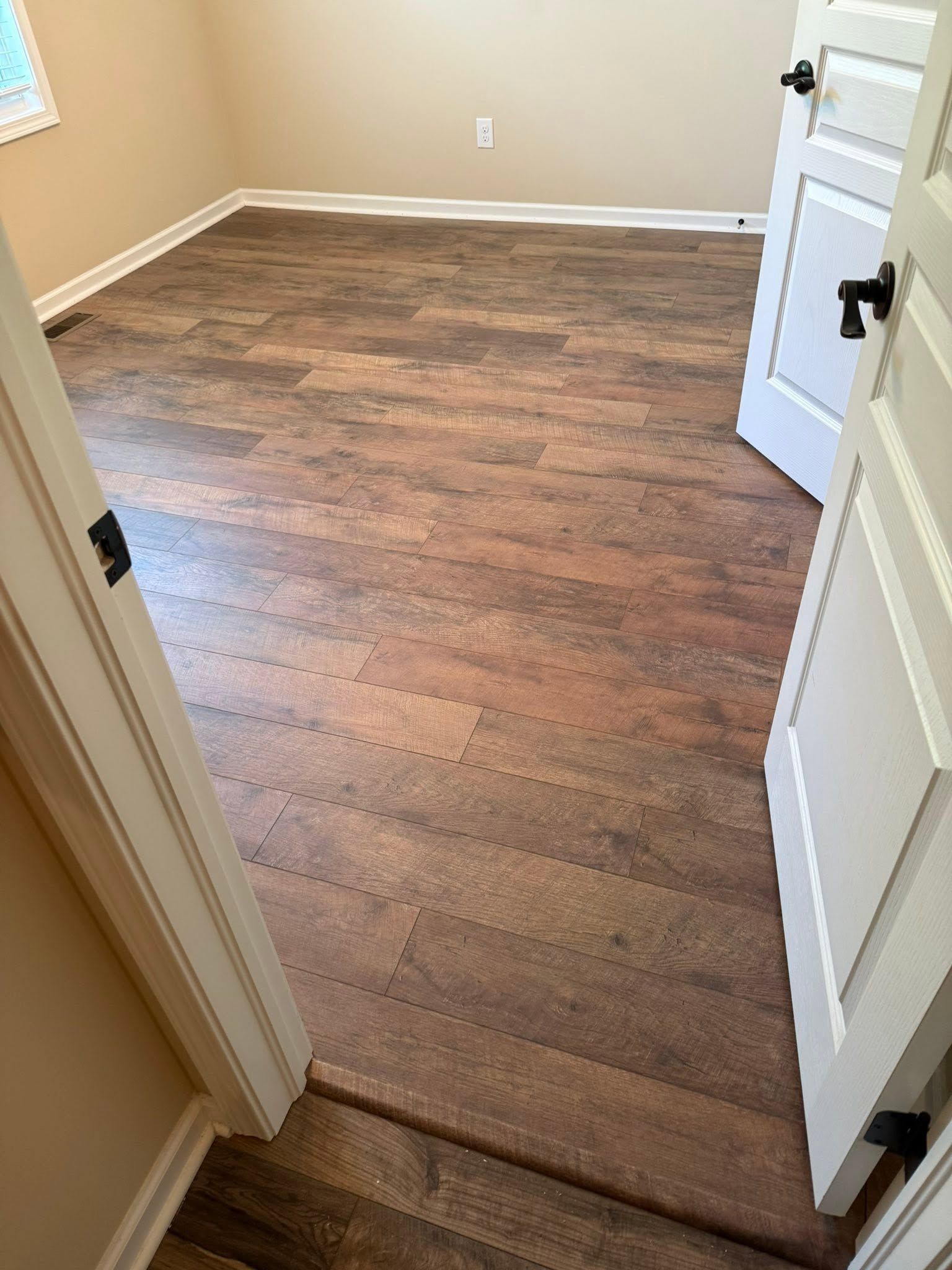 Empty beige room with wood-look flooring and a white door open in the foreground.