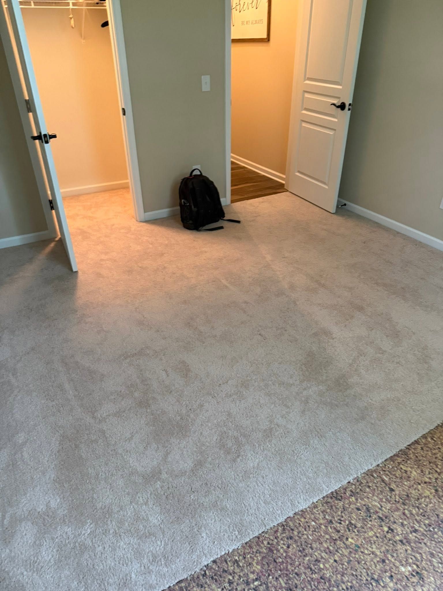 Empty carpeted room with beige walls, open doorways, and a black backpack on the floor.