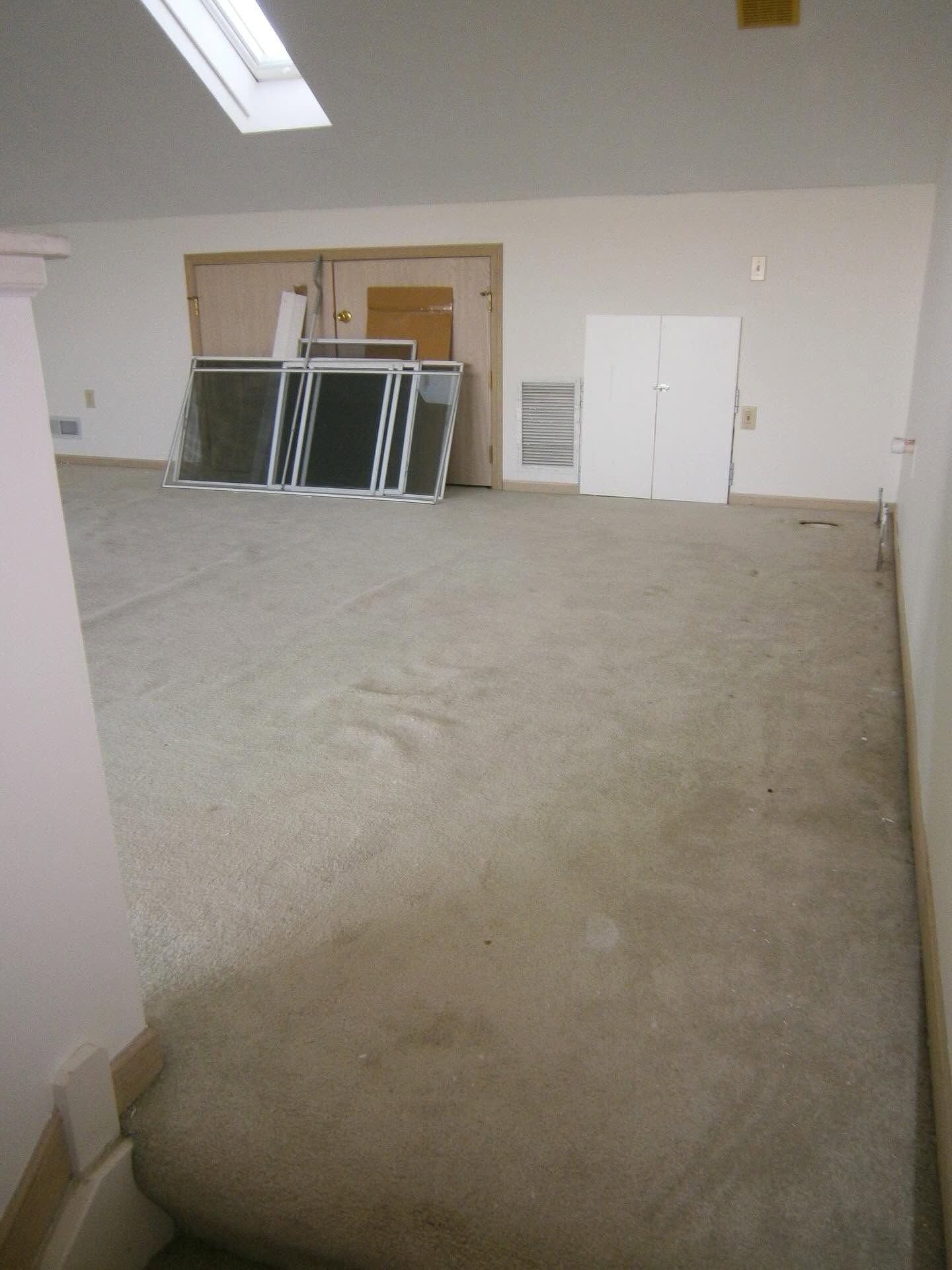 Empty beige room with carpeted floor, stacked window frames, and double doors on the back wall
