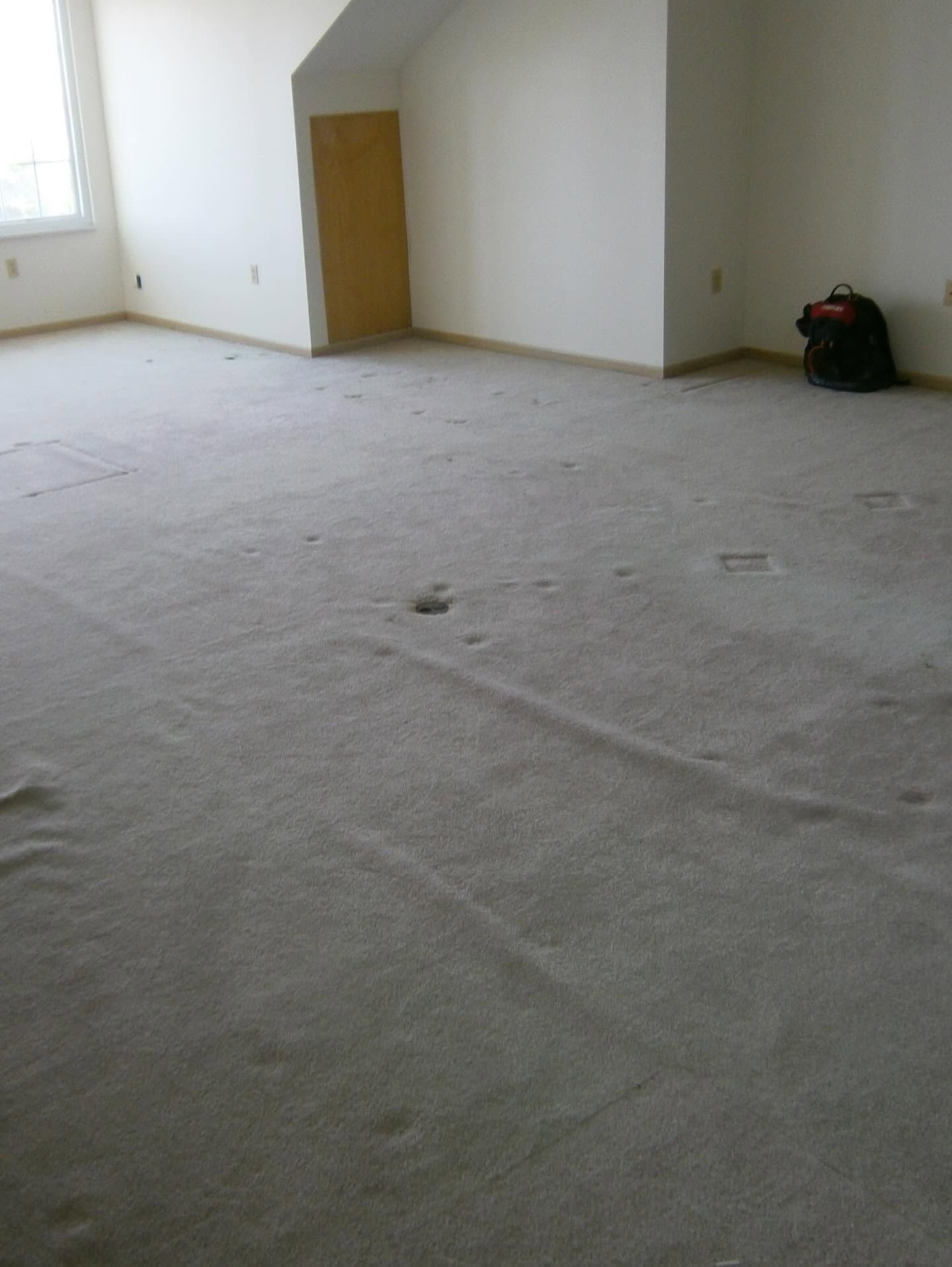 Empty room with unfinished carpet, white walls, a wooden door, and a bag in the corner.