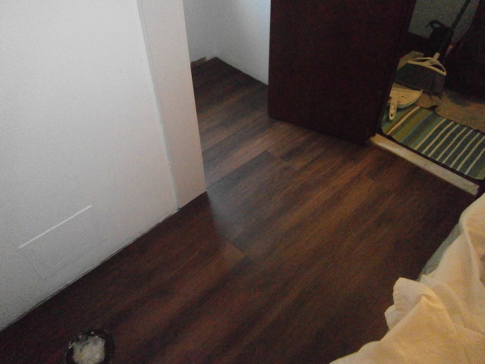 Empty room with dark wood floor, white walls, open doorway, and a bed edge in the foreground