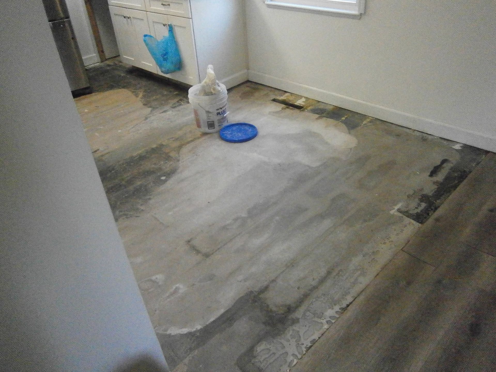 Partially stripped floor in a room with cleaning supplies and a blue bag near the wall
