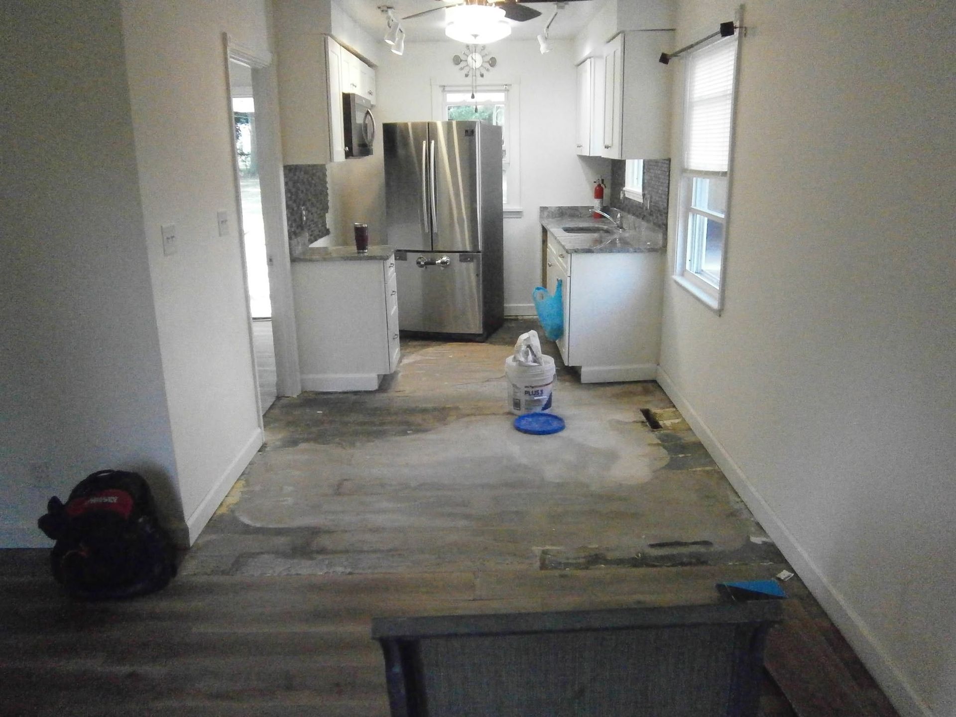 Empty hallway with worn flooring leading into a kitchen, with a bag on the left and cleaning items on the floor.