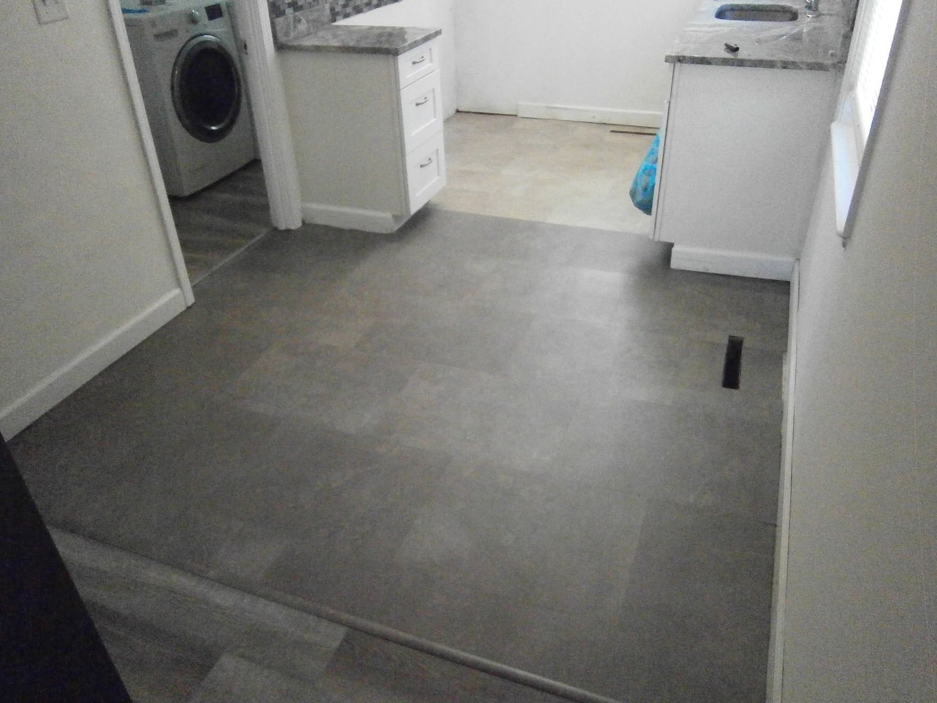 Empty laundry room with gray tile floor, washer, sink, and cabinets near a window.