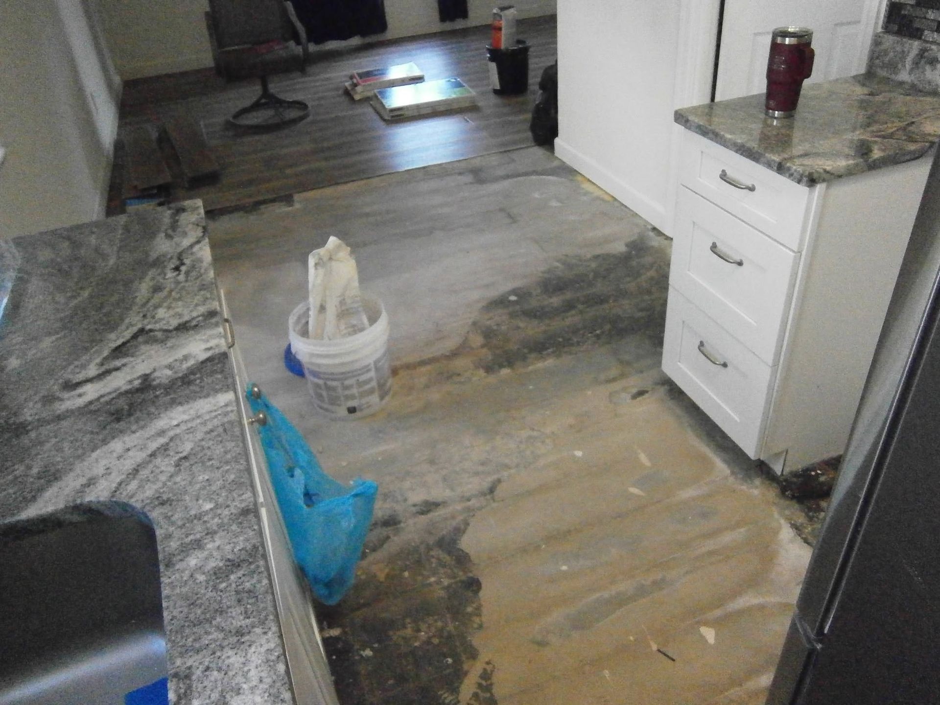 Messy kitchen floor with water damage, a mop bucket, and cleaning supplies beside cabinets and a countertop