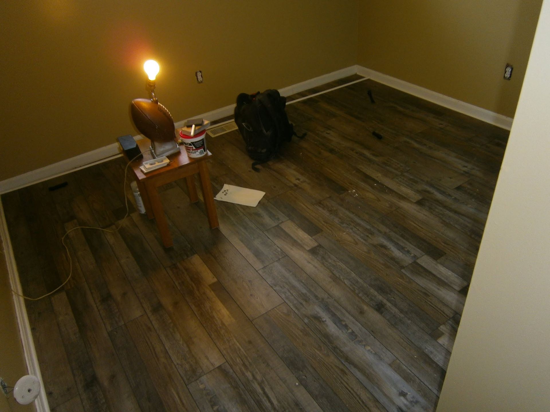 Empty room with wood flooring, a lit lamp on a small table, and a black bag near the wall.