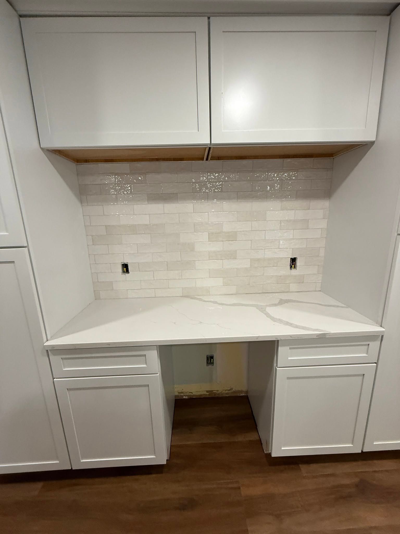White built-in desk nook with upper cabinets, white backsplash, and wood floor; drawers and open knee space below.