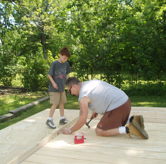 Two people painting a wooden deck outdoors, with a red paint tray nearby.