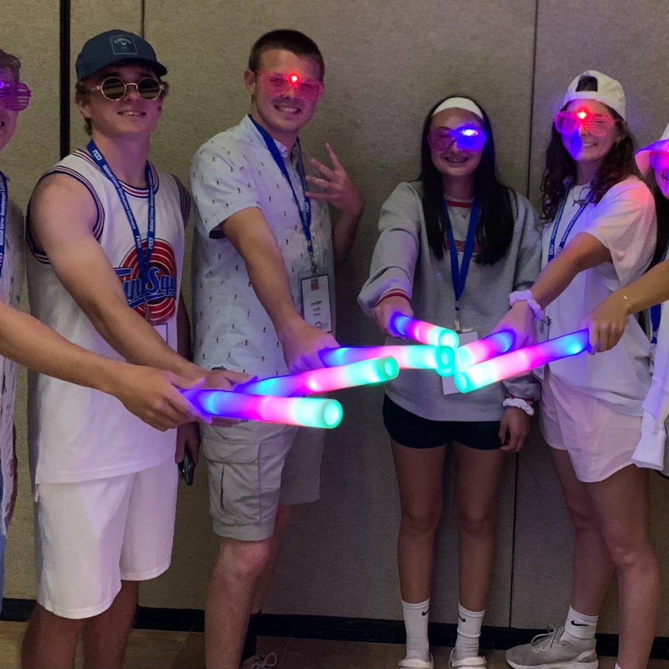 Group holding glowing LED batons in a hallway, smiling and posing in casual white outfits and party gear