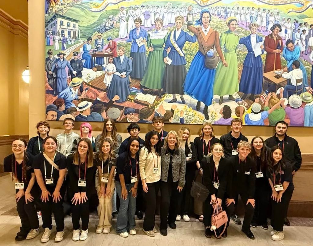 Group of people posing indoors in front of a colorful mural, with a large painted crowd scene behind them.