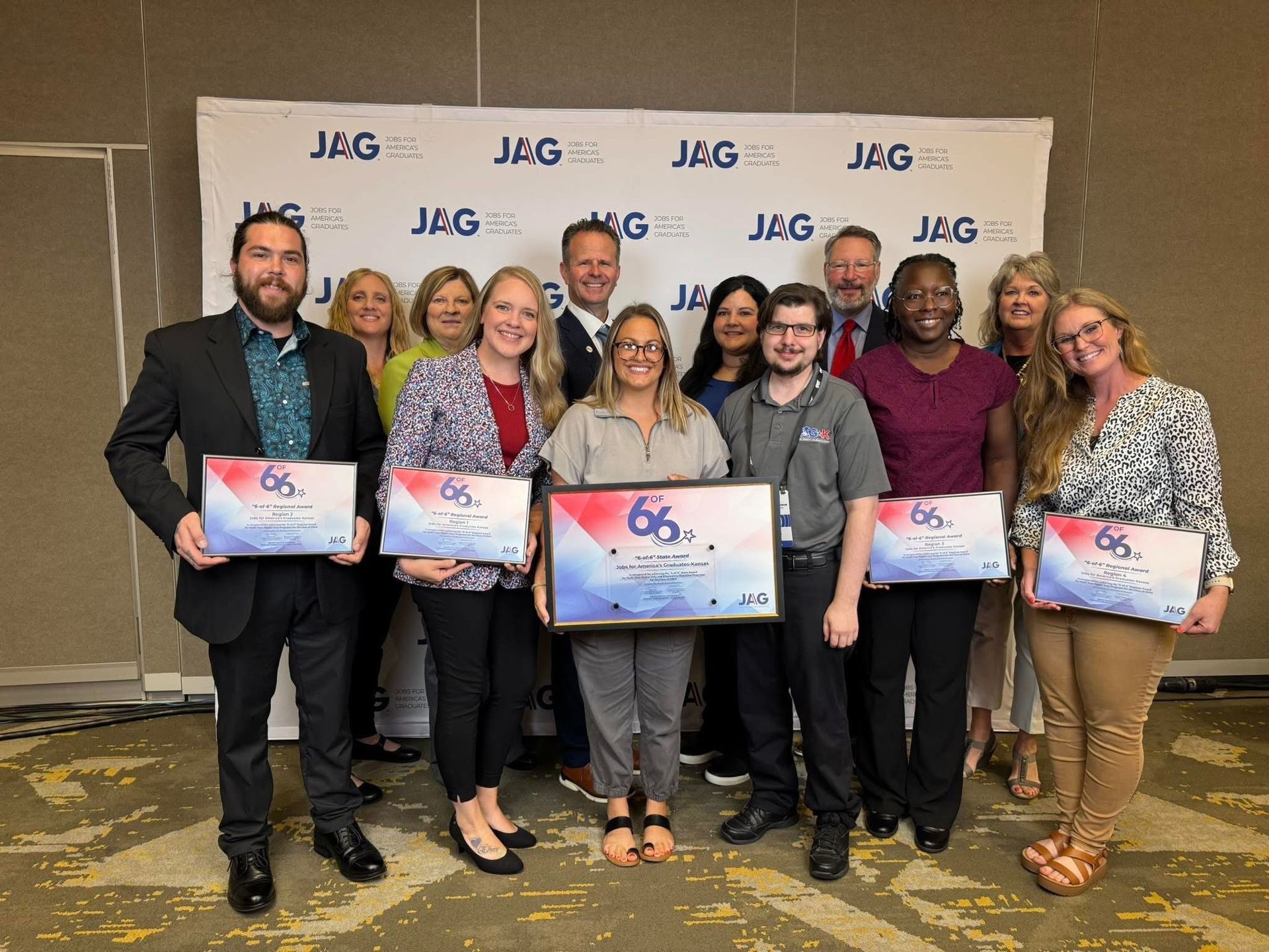 Group of people posing at a JAG event, holding framed certificates in front of a branded backdrop.