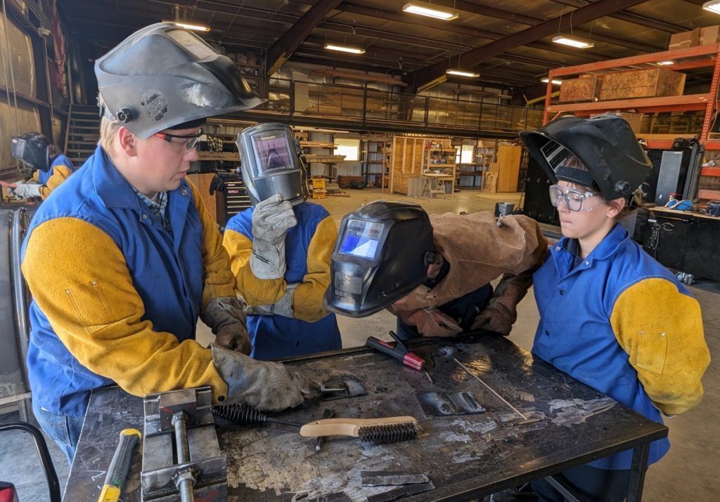Three welders in blue shirts and protective helmets work around a metal table in an industrial shop.