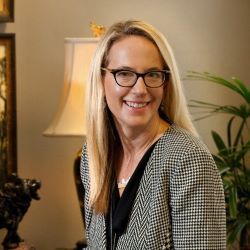 Smiling blonde woman wearing glasses and a checkered top in a warmly lit indoor room