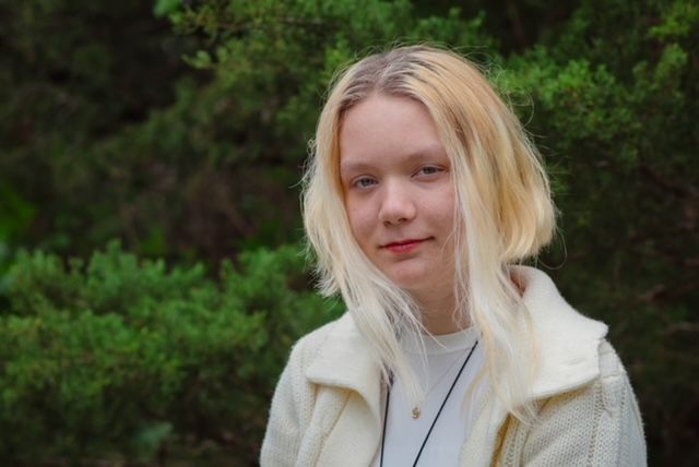 Person with blonde hair and white jacket standing outdoors against green foliage