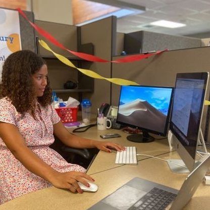 Person using a desktop computer at an office cubicle with a laptop and monitor on the desk.