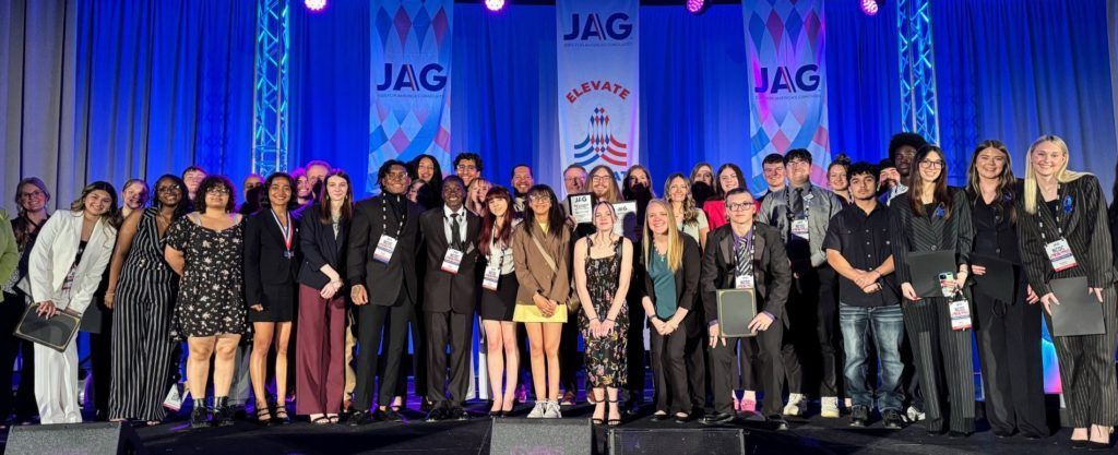 Group of people posing on a stage at an awards ceremony with blue curtains and JAG banners behind them