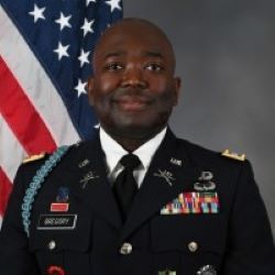 U.S. Army officer in dress uniform standing before an American flag, facing the camera with a slight smile