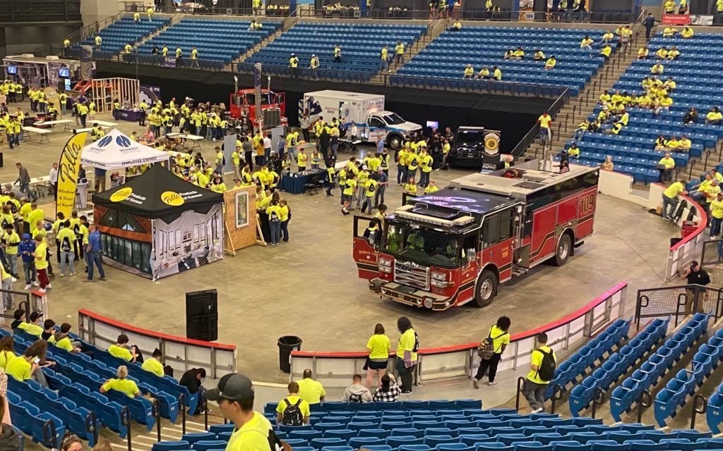 Indoors, a firetruck competition setup in an arena with people, barriers, and blue stadium seats.