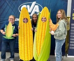 People posing with oversized corn props in front of a blue backdrop, smiling at the camera.