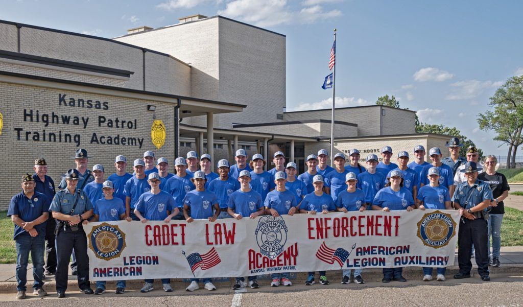 Group of Kansas Highway Patrol trainees in blue uniforms posing outside with a banner reading “Cadet Law Enforcement”