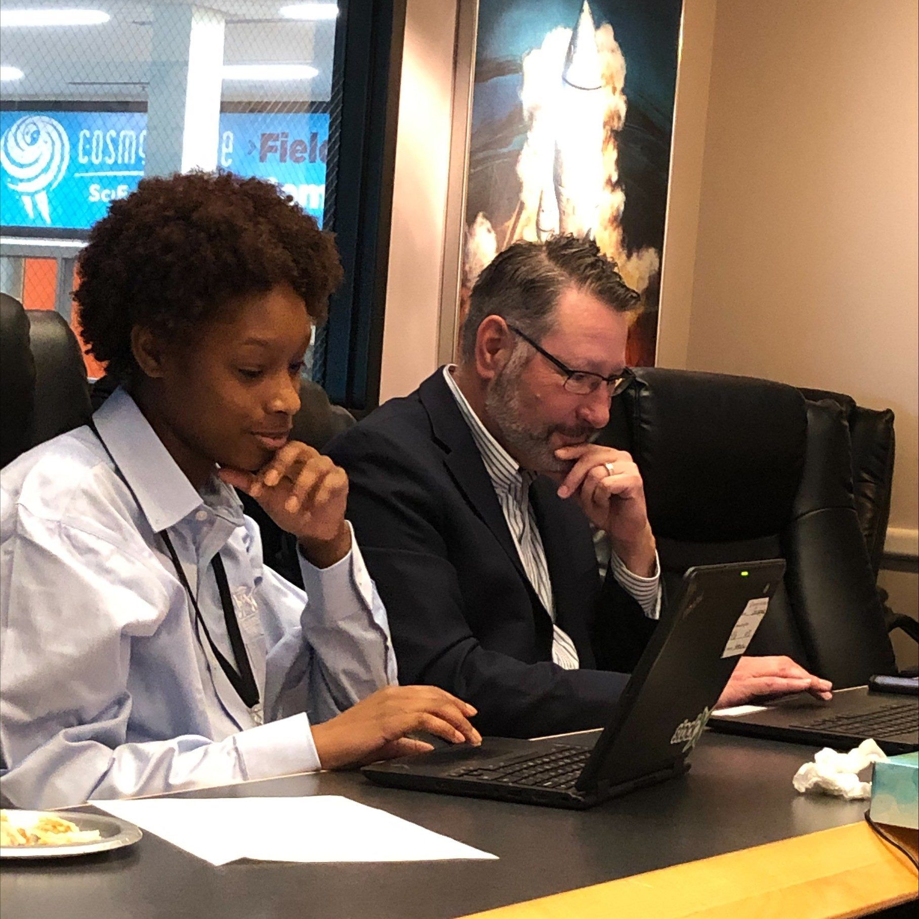 Two people in business attire working on laptops at a conference table in an office setting