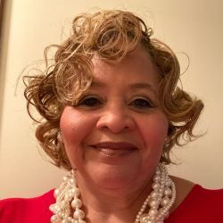 Smiling woman with curly blonde hair wearing a red top and pearl necklace against a beige wall