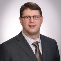 Professional headshot of a man in a dark suit, tie, and glasses, smiling against a light background