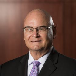 Professional headshot of a bald man in glasses and a black suit with a purple tie, smiling against a brown background