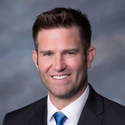 Professional headshot of a smiling man in a dark suit and blue tie against a gray background