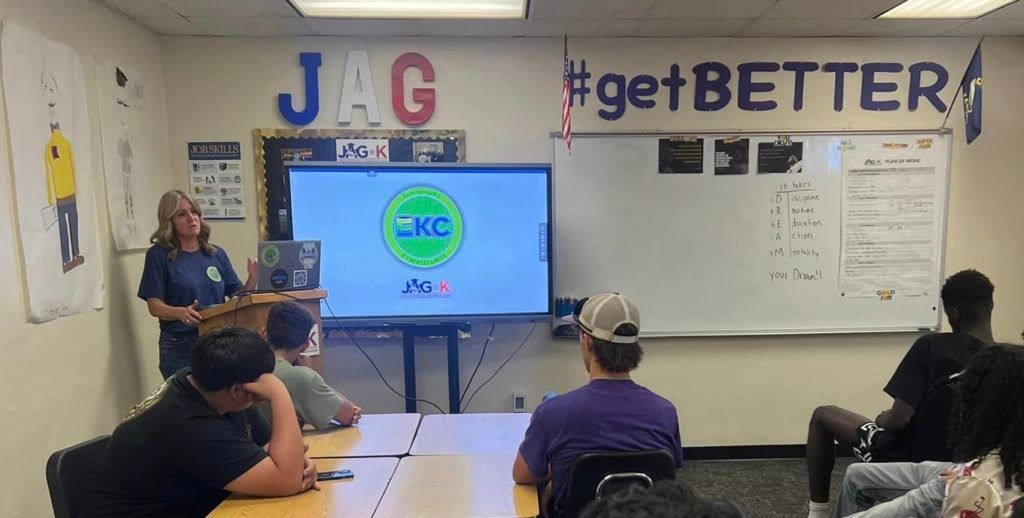 Classroom presentation with teacher beside a large screen showing a logo, students seated facing the front.