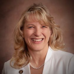 Smiling woman with blonde hair in a white blazer and pearl necklace against a warm studio backdrop