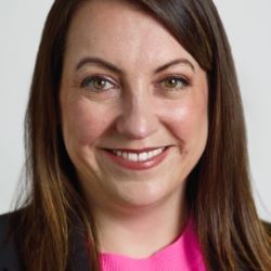 Close-up portrait of a smiling woman with brown hair, wearing a bright pink top, against a light background