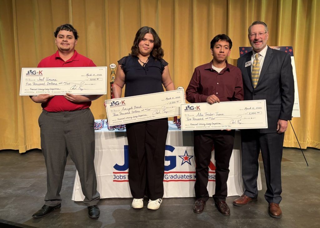 Three people holding oversized checks on a stage, with a man in a suit standing beside them.
