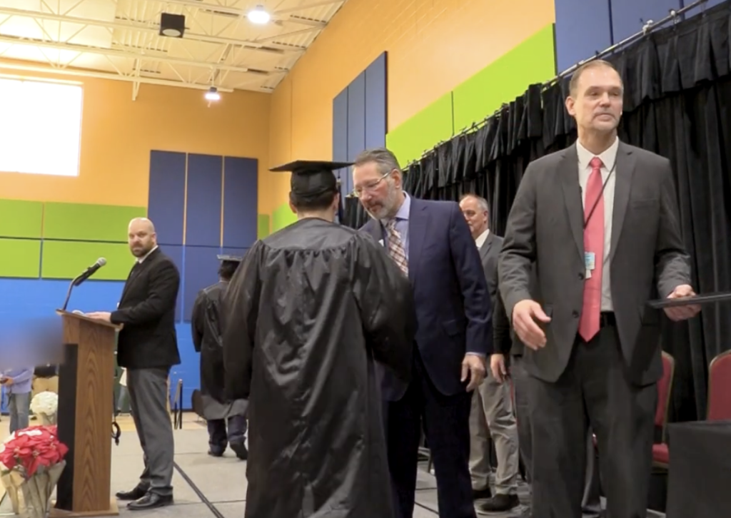 Graduation ceremony on a gym stage, with a graduate in cap and gown receiving congratulations from officials.