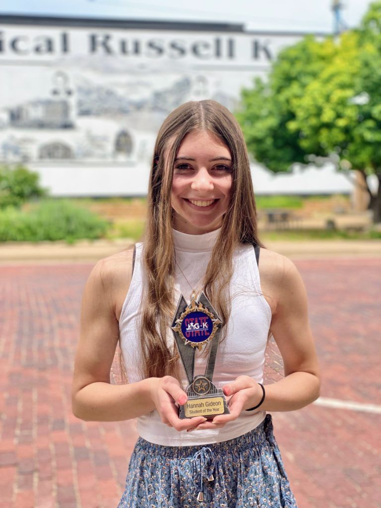 Smiling young woman holding a trophy outdoors in front of a campus building.