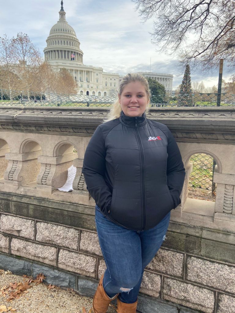 Person standing by a stone wall with the U.S. Capitol in the background, wearing a black jacket and jeans.
