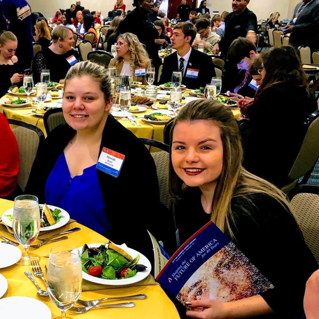 Two smiling attendees seated at a banquet table with plates of salad and drinks, in a crowded event hall