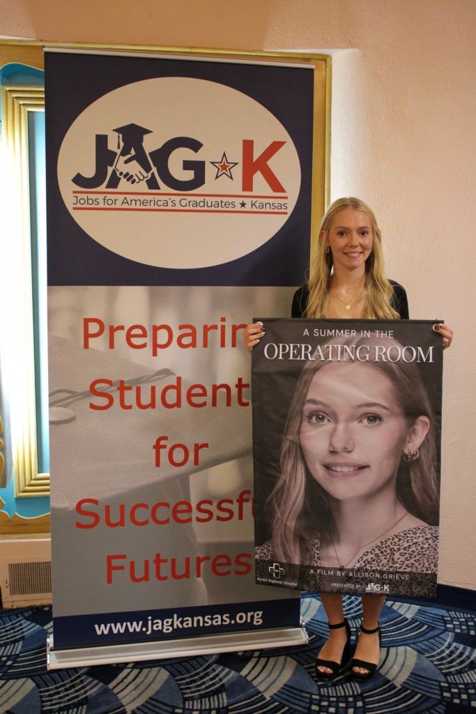 Woman holding a portrait poster beside a JAG-K banner at an indoor event