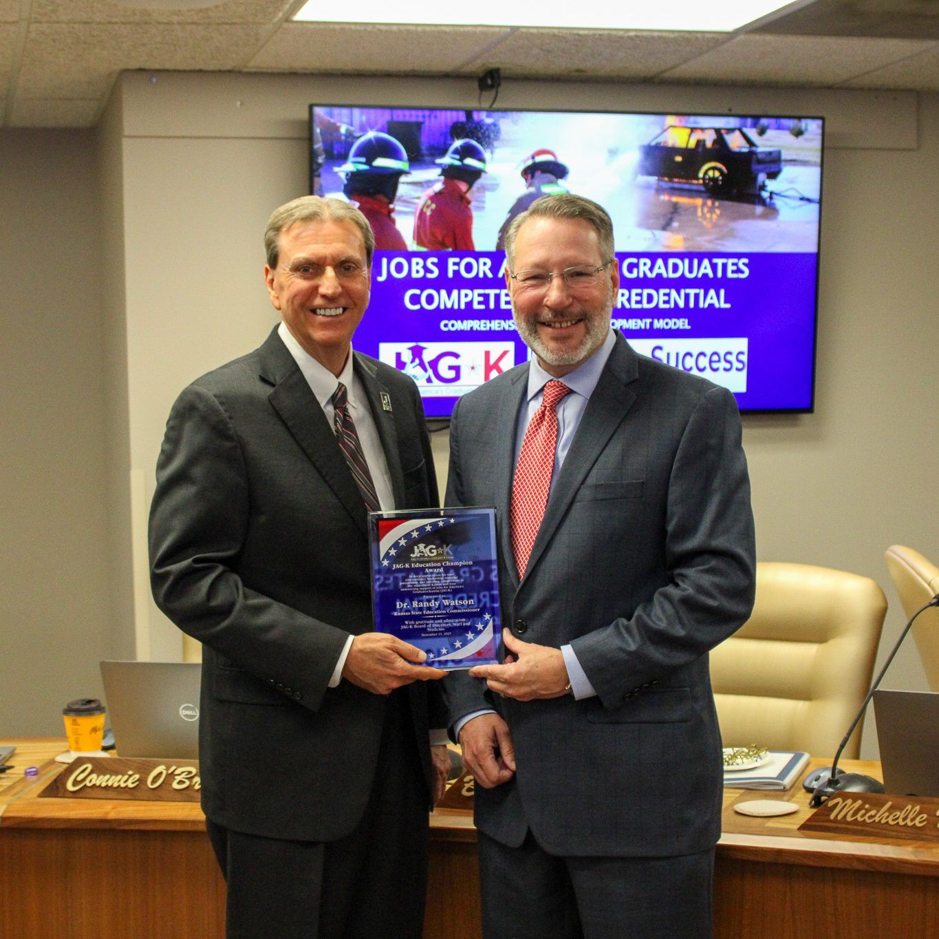 Two men in suits hold an award plaque in a conference room, with a presentation screen behind them.