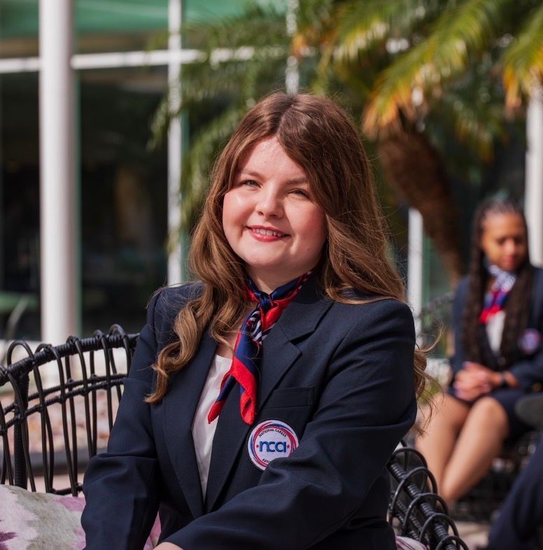 Woman in a navy blazer and red scarf sits smiling outdoors on a patio chair.