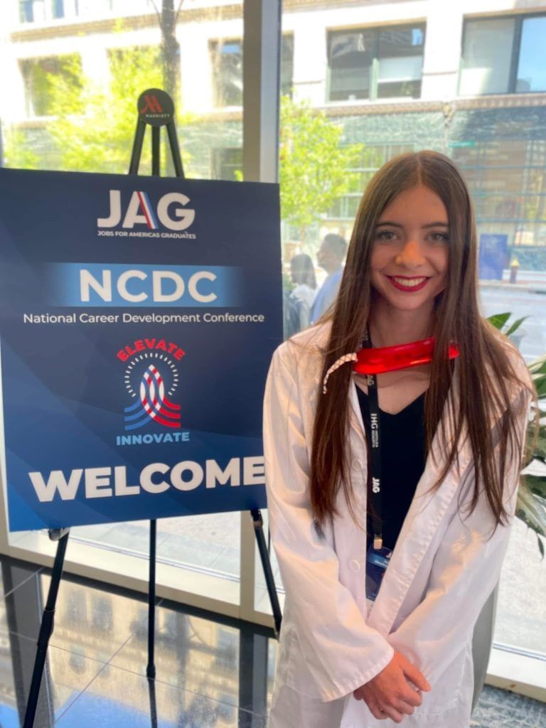 Woman standing beside a JAG NCDC welcome sign at a conference entrance.