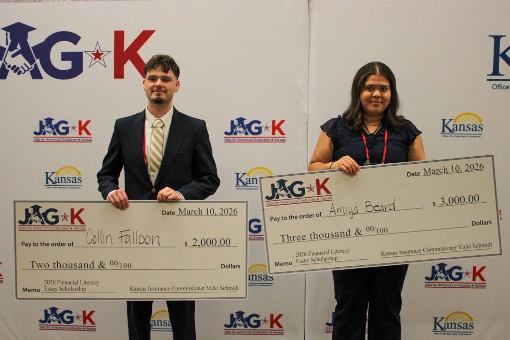 Two people on stage holding oversized checks at a JAG-K awards ceremony backdrop