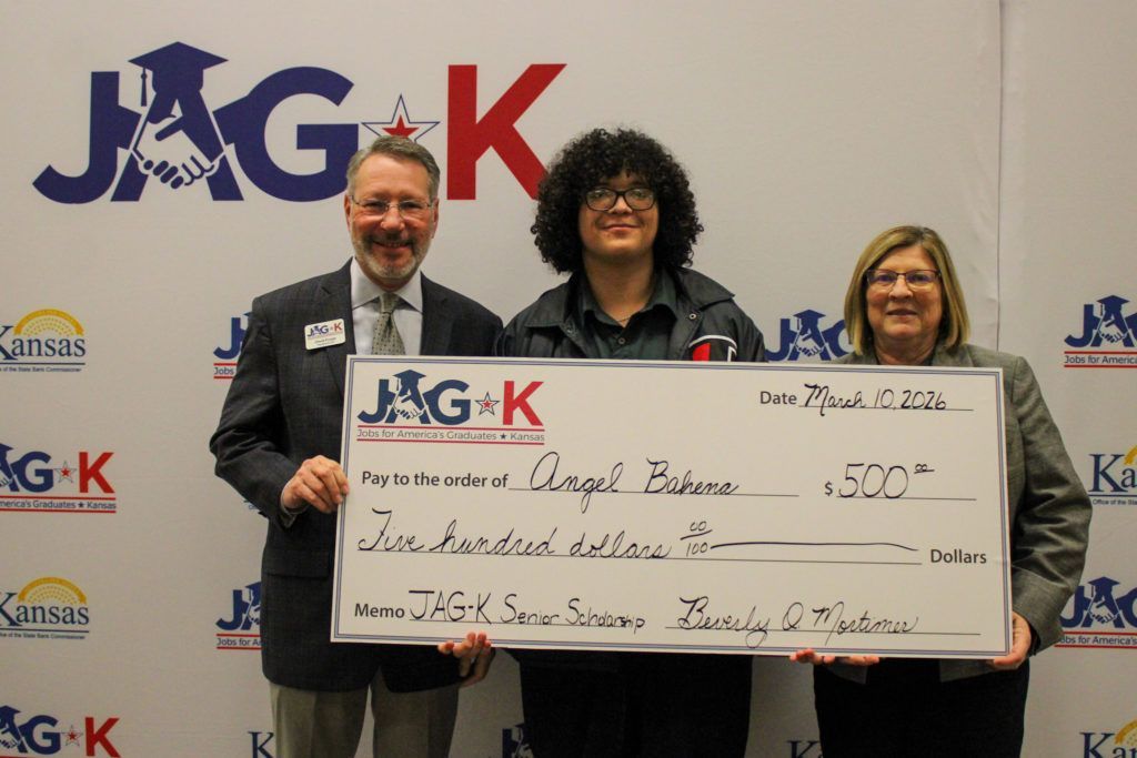 Three people holding a large ceremonial check at a JAG-K event backdrop