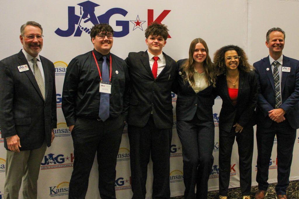 Six people in suits stand posing in front of a JAG-K banner at an event.