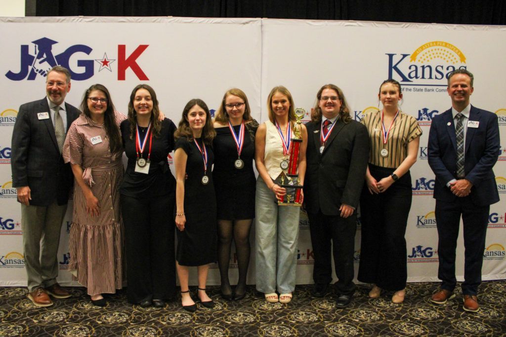 Nine adults posing on a conference stage in front of a JAG-K and Kansas backdrop