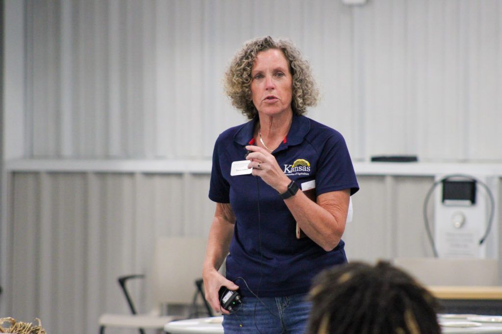 Person speaking indoors in a navy shirt, gesturing with one finger raised during a presentation.