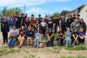 Large group posing outdoors in front of houses on a sunny day, with a blue sky and dirt foreground.